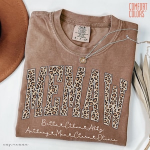 May include: Espresso-colored t-shirt with the word "MEMAW" in large leopard print letters. Below the word are names in cursive script. A silver necklace with a pendant is draped across the shirt.