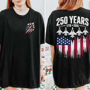 May include: A black short-sleeve t-shirt featuring a small graphic on the front left chest of a distressed American flag with fighter jets. The back displays the text 250 YEARS 1776-2026 above five fighter jets and a large distressed American flag.