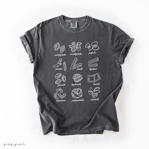 Op de afbeelding: Een donkergrijs T-shirt met een witte afbeelding van verschillende soorten pasta met hun namen onder elke afbeelding. De soorten pasta zijn: farfalle, conchiglie, pappardelle, cavatappi, penne, rigatoni, tagliatelle, gnocchi, maccheroni en ravioli.