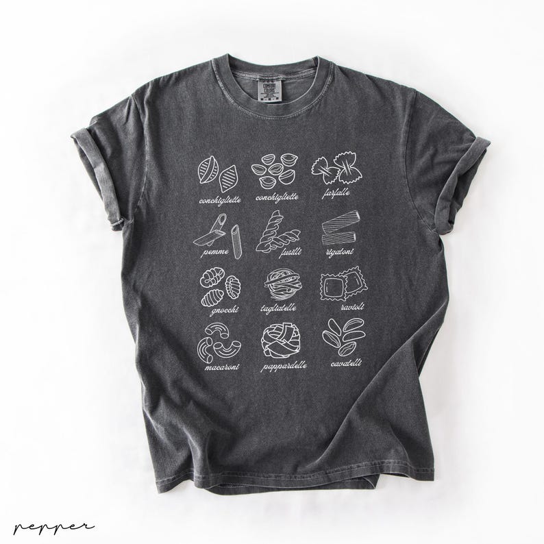 Op de afbeelding: Een donkergrijs T-shirt met een witte afbeelding van verschillende soorten pasta met hun namen onder elke afbeelding. De soorten pasta zijn: farfalle, conchiglie, pappardelle, cavatappi, penne, rigatoni, tagliatelle, gnocchi, maccheroni en ravioli.