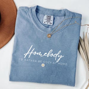 May include: A light blue t-shirt with the text "Homebody I'd rather be cozy at home." in white lettering. A small white daisy is below the text.