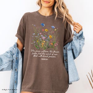 May include: Brown t-shirt with a floral design featuring blue, yellow, and pink flowers. The text reads "The grass withers, the flower fades, but the word of our God will stand forever. Isaiah 40:8."