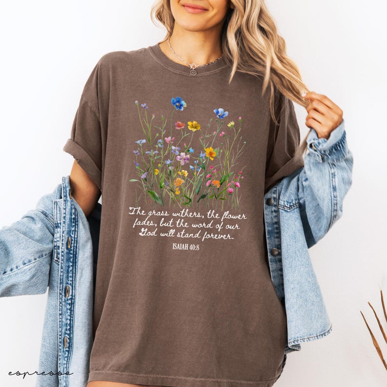 May include: Brown t-shirt with a floral design featuring blue, yellow, and pink flowers. The text reads "The grass withers, the flower fades, but the word of our God will stand forever. Isaiah 40:8."