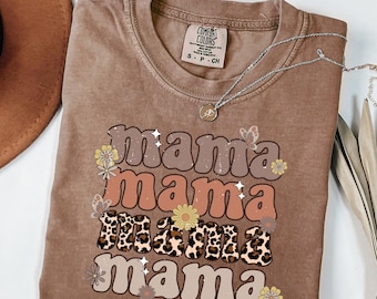 May include: A brown t-shirt with the text "mama mama mama mama" in a retro font with floral and leopard print accents. The shirt is made by Comfort Colors.