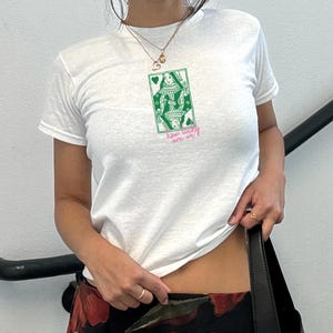 Puede incluir: Camiseta blanca con un dise&ntilde;o de carta de juego de la Reina de Corazones verde y blanco. El texto "how lucky are we" est&aacute; impreso en rosa debajo. La camiseta es de manga corta.