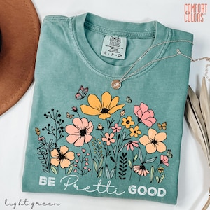 May include: Light green Comfort Colors t-shirt with a floral design featuring yellow and pink flowers, butterflies, and the phrase "BE Pretti GOOD." A silver necklace with a pendant is draped across the shirt.