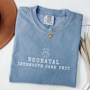 May include: A light blue t-shirt with a white outline of a teddy bear and the text "Neonatal Intensive Care Unit".