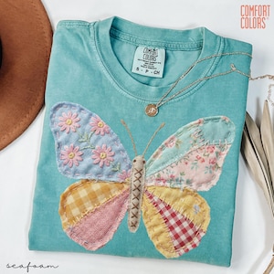 May include: Seafoam green Comfort Colors t-shirt with a large, patchwork butterfly design. The butterfly wings feature floral and plaid patterns in pink, yellow, and blue. A gold necklace with a pendant rests on the shirt. The shirt has the Comfort Colors label.