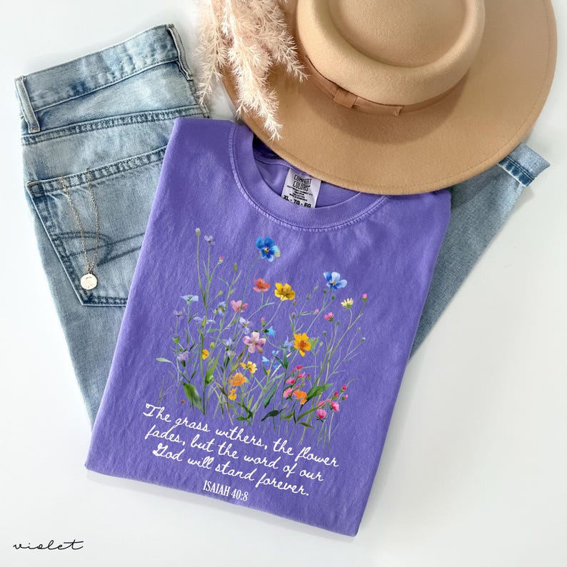 May include: A violet-colored t-shirt with a floral design and the text "The grass withers, the flower fades, but the word of our God will stand forever. ISAIAH 40:8." The shirt is paired with blue jeans and a tan hat.
