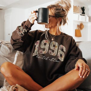 May include: A dark brown sweatshirt with the year "1996" in a floral design. The words "limited edition" are below the numbers. The sleeve has the text "Style Ten" in a heart shape. A black mug is held up.