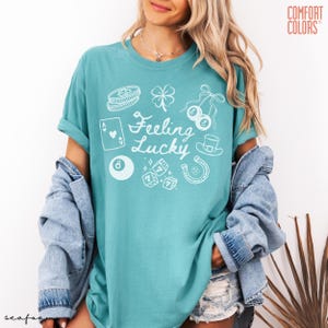 Może przedstawiać: Turkusowa koszulka Comfort Colors z napisem "Feeling Lucky" i białymi ilustracjami ikon o tematyce kasynowej. Projekt zawiera czterolistną koniczynę, kostki do gry, karty do gry i podkowę.