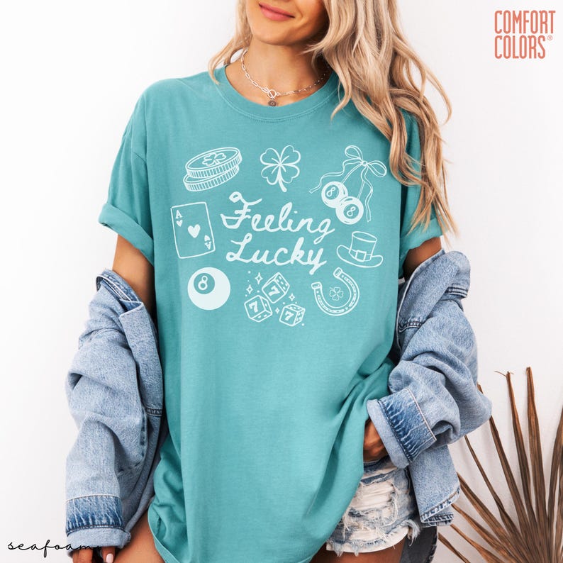 Może przedstawiać: Turkusowa koszulka Comfort Colors z napisem "Feeling Lucky" i białymi ilustracjami ikon o tematyce kasynowej. Projekt zawiera czterolistną koniczynę, kostki do gry, karty do gry i podkowę.