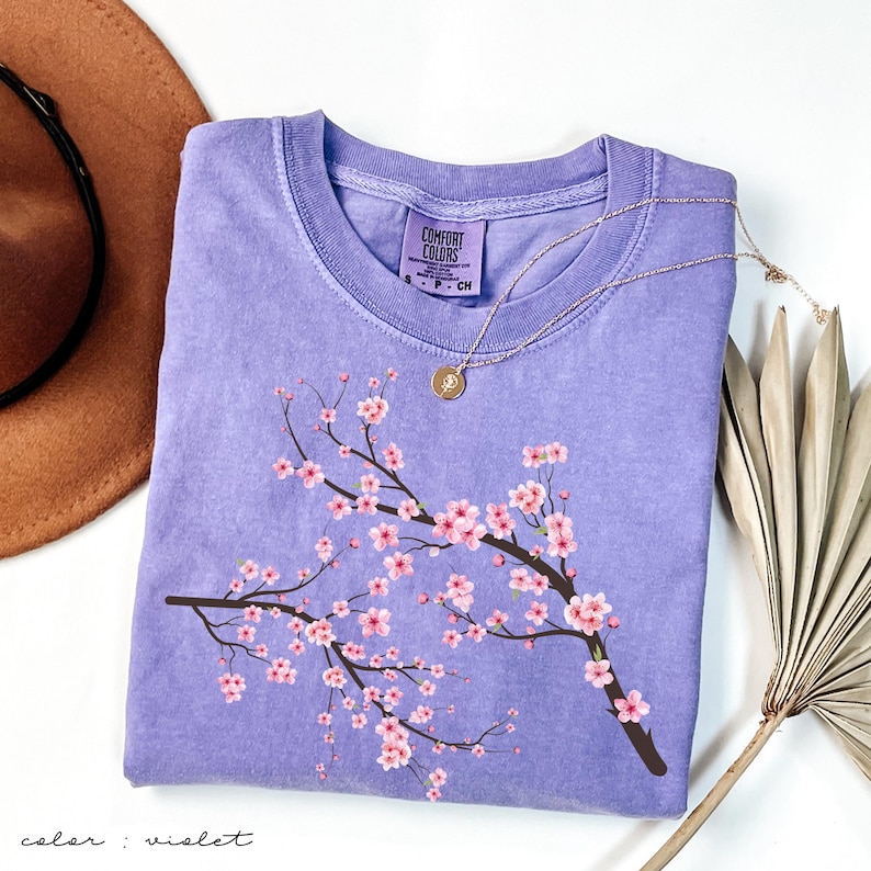 May include: A purple t-shirt with a pink cherry blossom branch design. The shirt has a crew neck and short sleeves. The text "Comfort Colors" is visible on the tag.