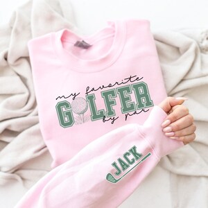 K&ouml;nnte beinhalten: Rosafarbener Pullover mit dem Text "my favorite golfer by par" und einer Golfballgrafik. Der &Auml;rmel hat den Namen "JACK" in gr&uuml;nem Text mit einer Golfclubgrafik.