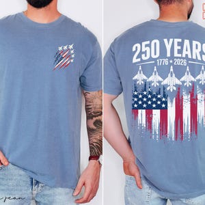 May include: A blue short-sleeve t-shirt featuring a small graphic on the front left chest of fighter jets over a distressed American flag. The back displays the text 250 YEARS 1776-2026 above five fighter jets and a large distressed American flag.