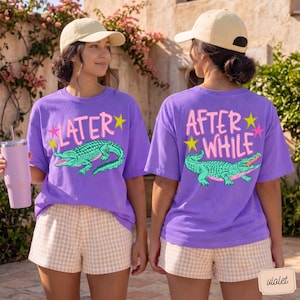 May include: Two women wearing violet t-shirts with a green alligator graphic. The front of the shirt says "LATER" and the back says "AFTER WHILE". They are also wearing tan baseball caps and checkered shorts.