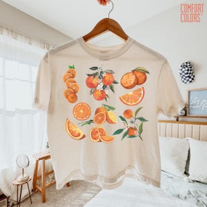 May include: A cream-colored t-shirt featuring a vibrant orange fruit design. The graphic includes whole oranges, slices, and orange blossoms. The shirt is hanging on a wooden hanger. The words "COMFORT COLORS" are printed on the upper right.