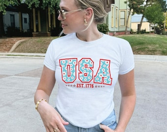 May include: White t-shirt with the word "USA" in red outlined letters filled with a blue floral pattern. Below the word "USA" is the text "EST. 1776" with stars. The shirt is short-sleeved.