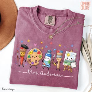 May include: A berry-colored Comfort Colors t-shirt featuring a graphic of anthropomorphic art supplies including a paintbrush, palette, paint tube, and easel. The text "Mrs. Anderson" is printed in white script below the characters, with small stars and hearts.