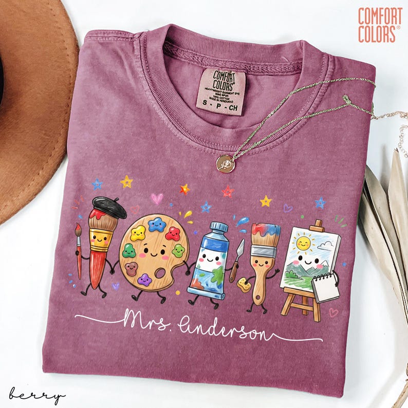 May include: A berry-colored Comfort Colors t-shirt featuring a graphic of anthropomorphic art supplies including a paintbrush, palette, paint tube, and easel. The text "Mrs. Anderson" is printed in white script below the characters, with small stars and hearts.