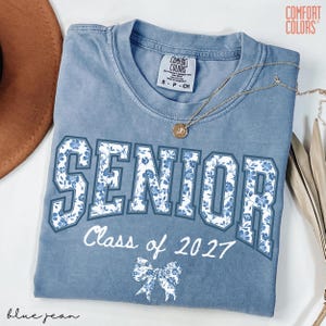 May include: A blue jean Comfort Colors t-shirt with the word "SENIOR" in large white letters filled with a blue floral pattern. Below, the text "Class of 2027" and a floral bow are printed in white. A gold necklace rests on the shirt.