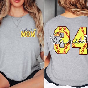 Puede incluir: Una camiseta gris claro con las palabras "Softball MOM" en amarillo y negro en la parte delantera. La parte trasera de la camiseta presenta el n&uacute;mero "84" en amarillo con un dise&ntilde;o de softbol y el nombre "David" en blanco.