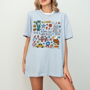 K&ouml;nnte beinhalten: Hellblaues T-Shirt mit einer farbenfrohen Grafik zum 250-j&auml;hrigen Jubil&auml;um Amerikas. Das Design zeigt die Freiheitsstatue, einen Baseball, das Kapitol, einen Adler und den Text "Happy 250 America 2026".
