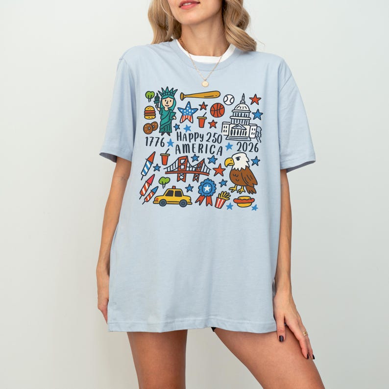 K&ouml;nnte beinhalten: Hellblaues T-Shirt mit einer farbenfrohen Grafik zum 250-j&auml;hrigen Jubil&auml;um Amerikas. Das Design zeigt die Freiheitsstatue, einen Baseball, das Kapitol, einen Adler und den Text "Happy 250 America 2026".