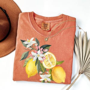 May include: Coral-colored Comfort Colors t-shirt featuring a watercolor design of lemons, white flowers, and green leaves. A gold necklace with a pendant rests on the shirt. A brown hat and dried palm leaf are in the background.
