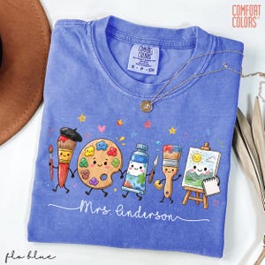 May include: A periwinkle blue Comfort Colors t-shirt featuring a graphic of cartoon art supplies including brushes, a palette, paint, and an easel with a sun drawing. The text Mrs. Linderson is printed in white script below the characters.