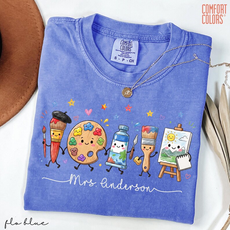 May include: A periwinkle blue Comfort Colors t-shirt featuring a graphic of cartoon art supplies including brushes, a palette, paint, and an easel with a sun drawing. The text Mrs. Linderson is printed in white script below the characters.