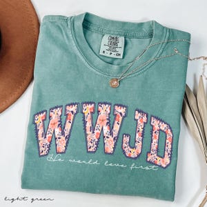 May include: Light green Comfort Colors t-shirt with the letters "WWJD" in a floral design. The text "He would love first" is below the letters. A gold necklace and a brown hat are also visible.
