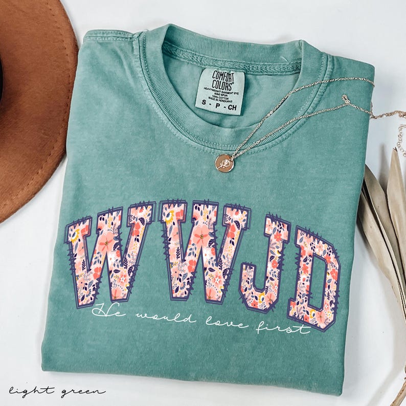May include: Light green Comfort Colors t-shirt with the letters "WWJD" in a floral design. The text "He would love first" is below the letters. A gold necklace and a brown hat are also visible.