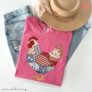 May include: A pink Comfort Colors t-shirt featuring a patchwork chicken design with red, blue, and white patterns. The image also includes a pair of blue jeans, a tan hat, and a necklace. The word "crunchberry" is written in cursive at the bottom.
