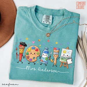 May include: A seafoam green t-shirt featuring a graphic of cartoon art supplies including a paintbrush, palette, paint tube, and easel with a sun painting. The text "Mrs. Linderson" is printed in white script below the characters, with "Comfort Colors" on the neck label.
