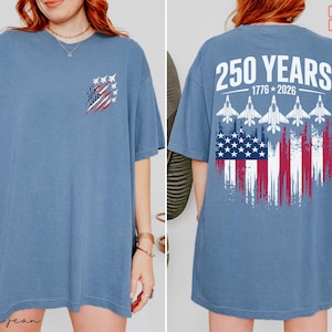 May include: A blue short-sleeve t-shirt featuring a small graphic on the front left chest of fighter jets and a flag. The back displays the text 250 YEARS 1776-2026 above five fighter jets and a distressed American flag graphic design.