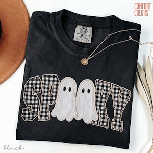 May include: A black short-sleeved t-shirt with a crew neckline featuring the word SPOOKY appliqued across the chest. The letters S, P, K, and Y are made of black and white buffalo plaid fabric, while two white ghost shapes replace the double O.