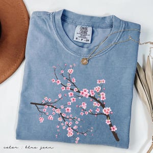 May include: A blue jean t-shirt with a pink cherry blossom branch design. The shirt has a crew neck and short sleeves. The text "Comfort Colors" is visible on the tag.