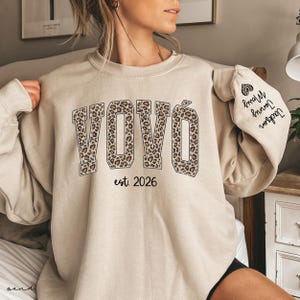 May include: Beige sweatshirt with the word "VOVÓ" in leopard print lettering and the text "est. 2026". The sleeve features a heart graphic and the names "Jackson, Jonny, Merry".