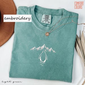 May include: Light green t-shirt with an embroidered penguin and mountain design. The shirt has a crew neck and short sleeves. A silver necklace with a pendant is on the shirt. The text "embroidery" is on the left side of the shirt.