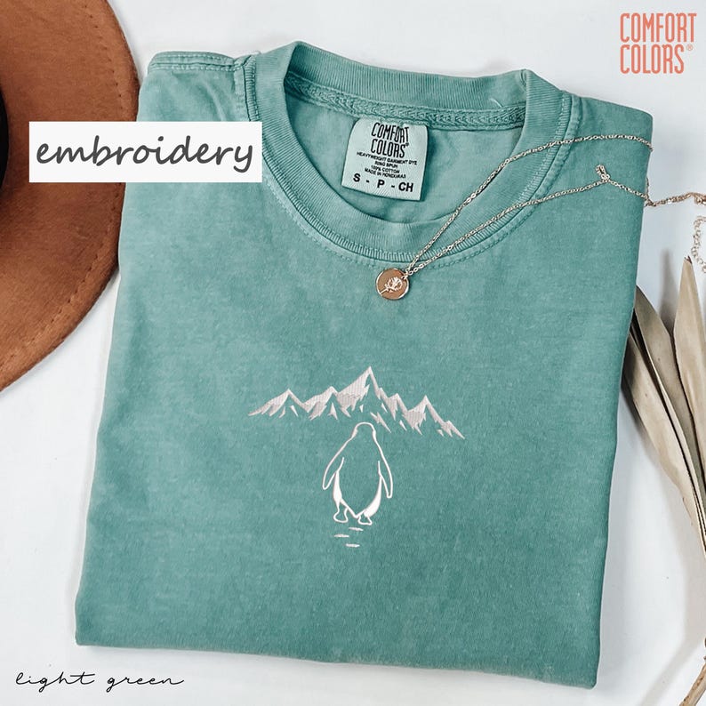 May include: Light green t-shirt with an embroidered penguin and mountain design. The shirt has a crew neck and short sleeves. A silver necklace with a pendant is on the shirt. The text "embroidery" is on the left side of the shirt.