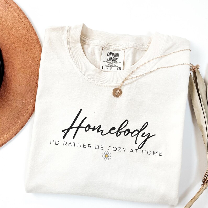 May include: A cream colored t-shirt with the text "Homebody I'd rather be cozy at home." in black lettering. A small white daisy is below the text.