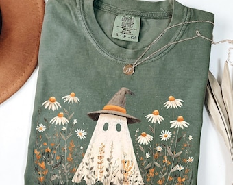 May include: Green Comfort Colors t-shirt featuring a ghost in a witch hat amidst daisies. A silver necklace with a pendant rests on the shirt. The shirt is size S-P-CH.