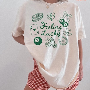 Może przedstawiać: Kremowa koszulka z zielonym nadrukiem przedstawiającym symbole szczęścia, takie jak czterolistna koniczyna, kostki do gry i podkowa. Tekst "Feeling Lucky" jest umieszczony na środku koszulki. Koszulka jest połączona z czerwono-białymi szortami w kratę.
