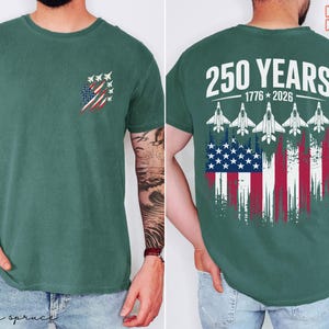 May include: A dark green t-shirt featuring a small graphic on the front left chest of fighter jets over a distressed American flag. The back displays the text 250 YEARS 1776-2026 above five fighter jets and a large distressed American flag graphic.