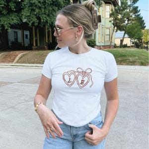 May include: White short-sleeved t-shirt with a red heart-shaped design. The design features two connected hearts with the letters "L" and "B" inside. The shirt is worn by a person wearing sunglasses and gold jewelry.