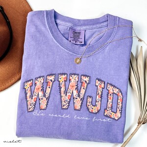 May include: A lilac Comfort Colors t-shirt featuring the letters WWJD in a floral pattern. The text "He would love first" is below the letters. A gold necklace and a brown hat are also in the image.