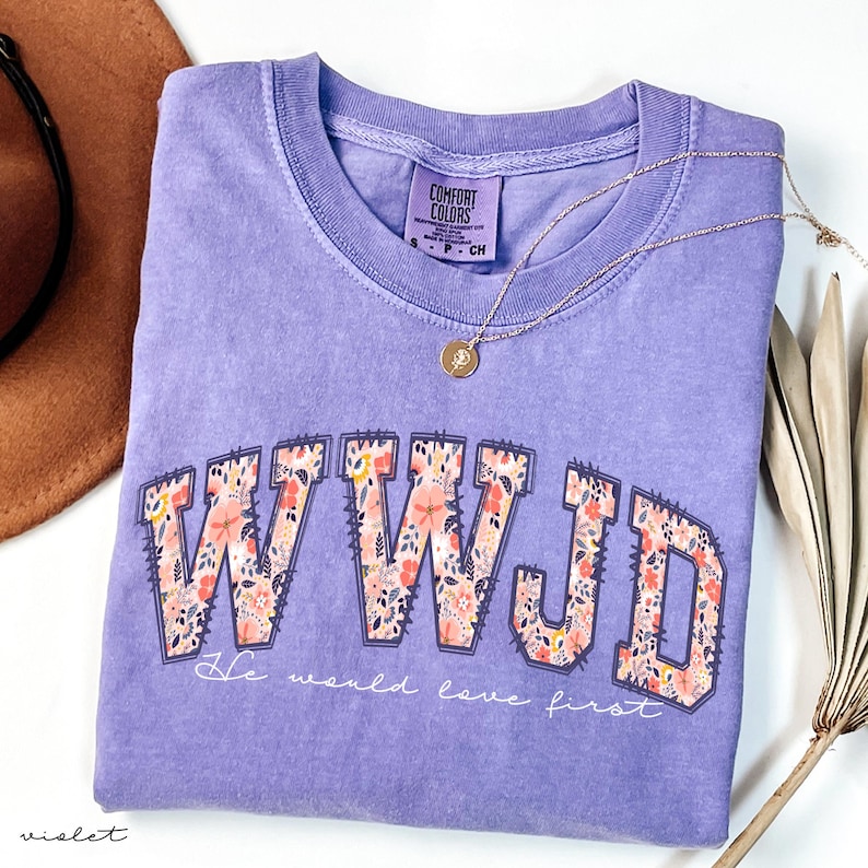 May include: A lilac Comfort Colors t-shirt featuring the letters WWJD in a floral pattern. The text "He would love first" is below the letters. A gold necklace and a brown hat are also in the image.