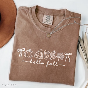 May include: A brown Comfort Colors t-shirt with the words "hello fall" and fall-themed graphics in white. The graphics include a bow, pumpkin, hat, coffee cup, scarf, leaf, and another bow. A silver necklace with a pendant is on the shirt.