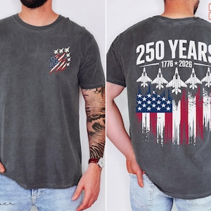 May include: A charcoal gray short-sleeve t-shirt featuring a small graphic on the front left chest of a stylized American flag with fighter jets. The back displays the text 250 YEARS 1776-2026 above five fighter jets and a distressed American flag.